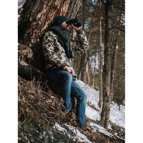 Ein Mann mit Fernglas sitzt im Winterwald neben einem Baum.
