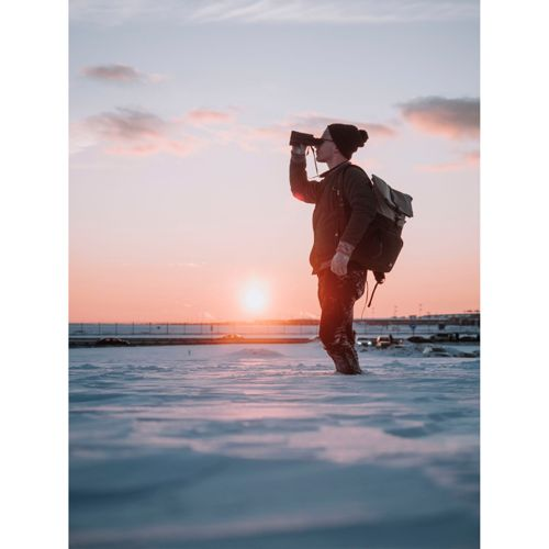 Ein Mann mit Rucksack schaut durch Fernglas in verschneiter Landschaft bei Sonnenuntergang.