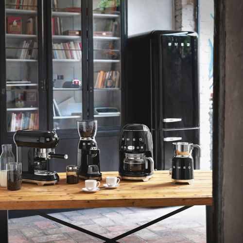 Machines à café et moulins SMEG noirs sur table en bois devant une bibliothèque.