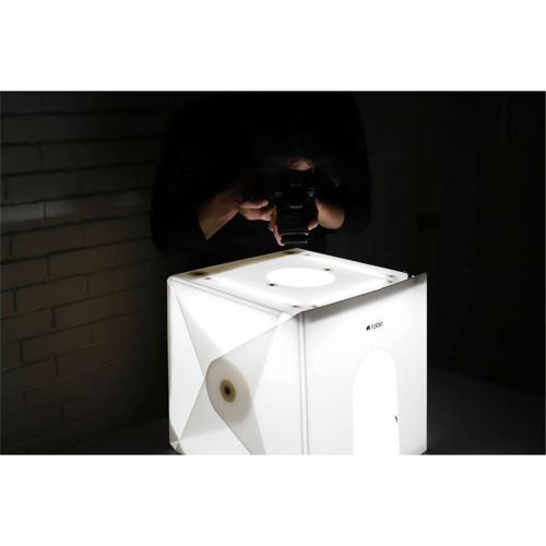 Eine Person fotografiert eine weiße Fotobox mit Lichtern. Ziegelwand im Hintergrund.