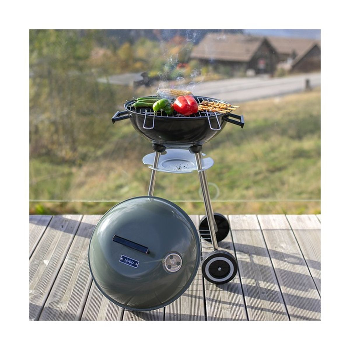 Ein Grill mit Gemüse und Spießen auf einer Holzterrasse, im Hintergrund Häuser.