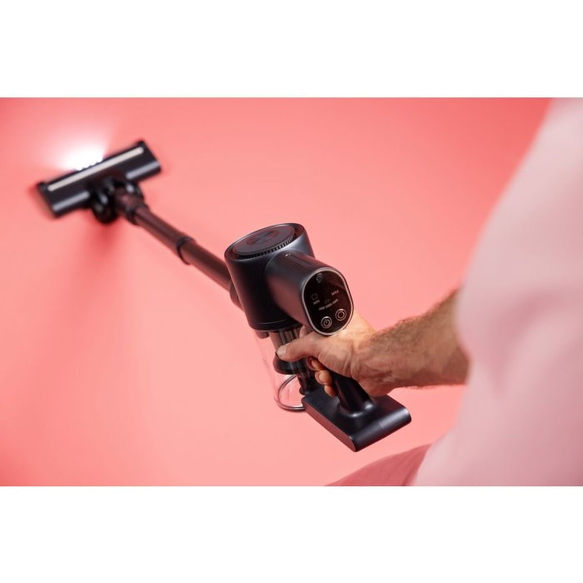 Een hand houdt een zwarte draadloze stofzuiger met licht op de borstelkop tegen een roze muur.
