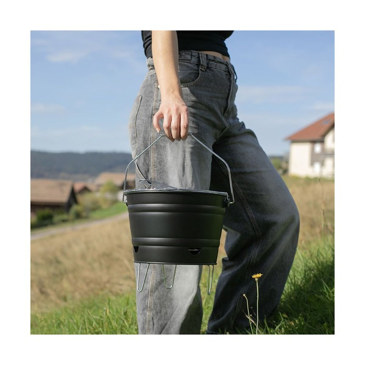 A person holds a black charcoal chimney starter with a metal handle outdoors.
