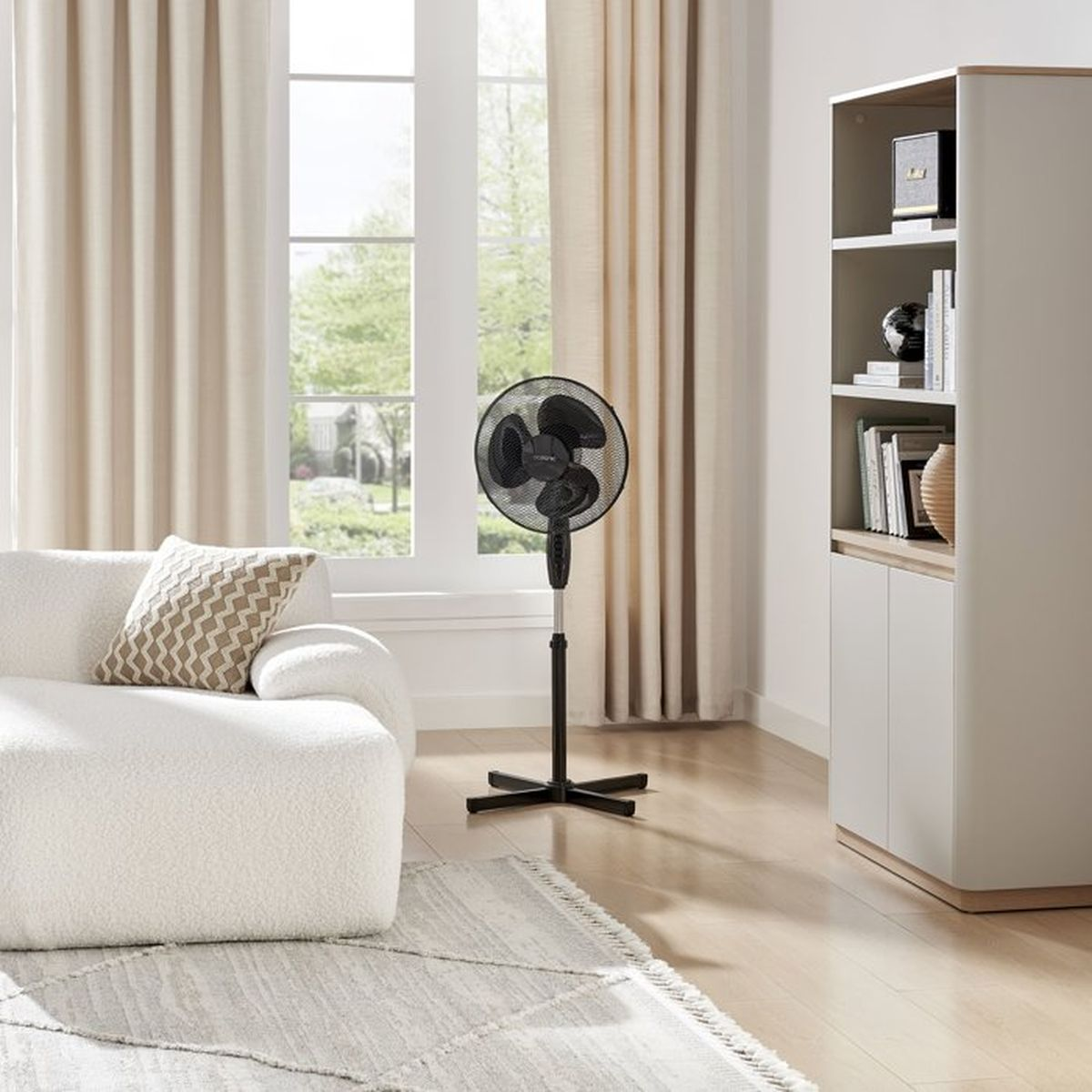 Ein schwarzer Standventilator steht in einem hellen Wohnzimmer neben einem weißen Sofa.