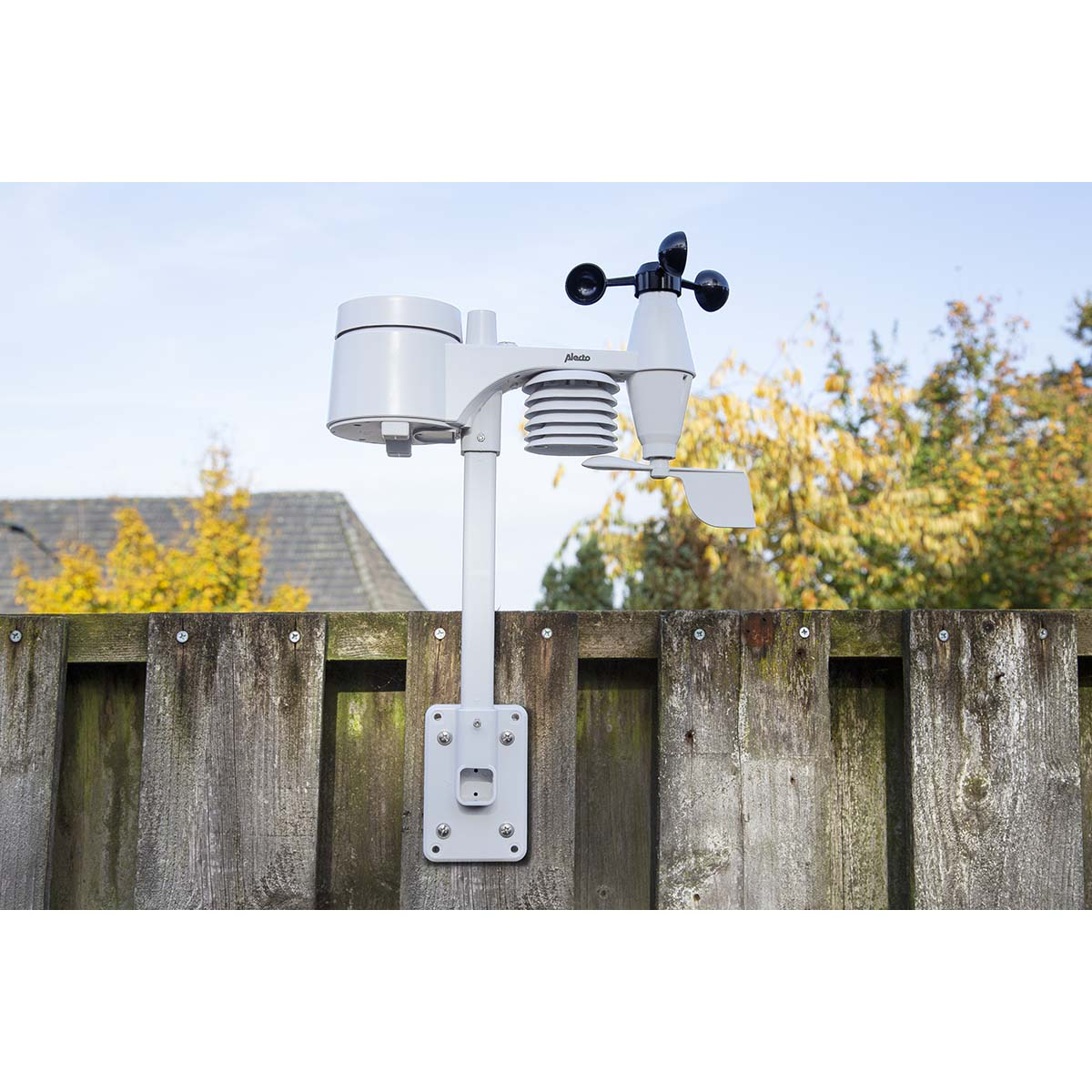 Een weerstation sensor gemonteerd op een houten schutting, met bomen op de achtergrond.