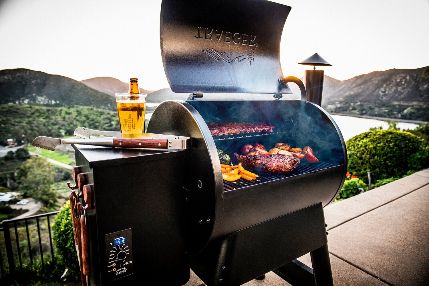 Un gril avec de la viande, des légumes et de la bière sur un patio avec des montagnes et de l'eau en arrière-plan.