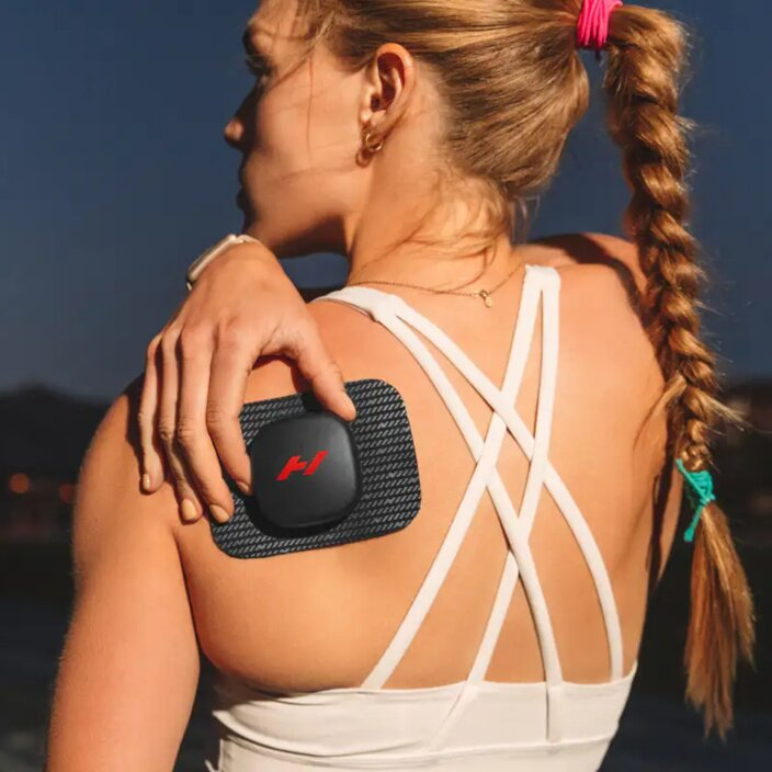 Frau mit schwarzen EMS-Gerät auf der Schulter, im Freien bei Dämmerung.