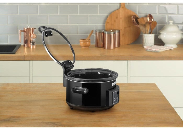 Una slow cooker nera con coperchio in vetro aperto su un tavolo di legno.