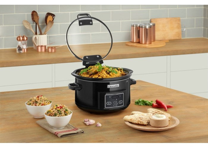 Una slow cooker nera con coperchio in vetro aperto, piena di cibo, accanto a ciotole di riso e pane pita.
