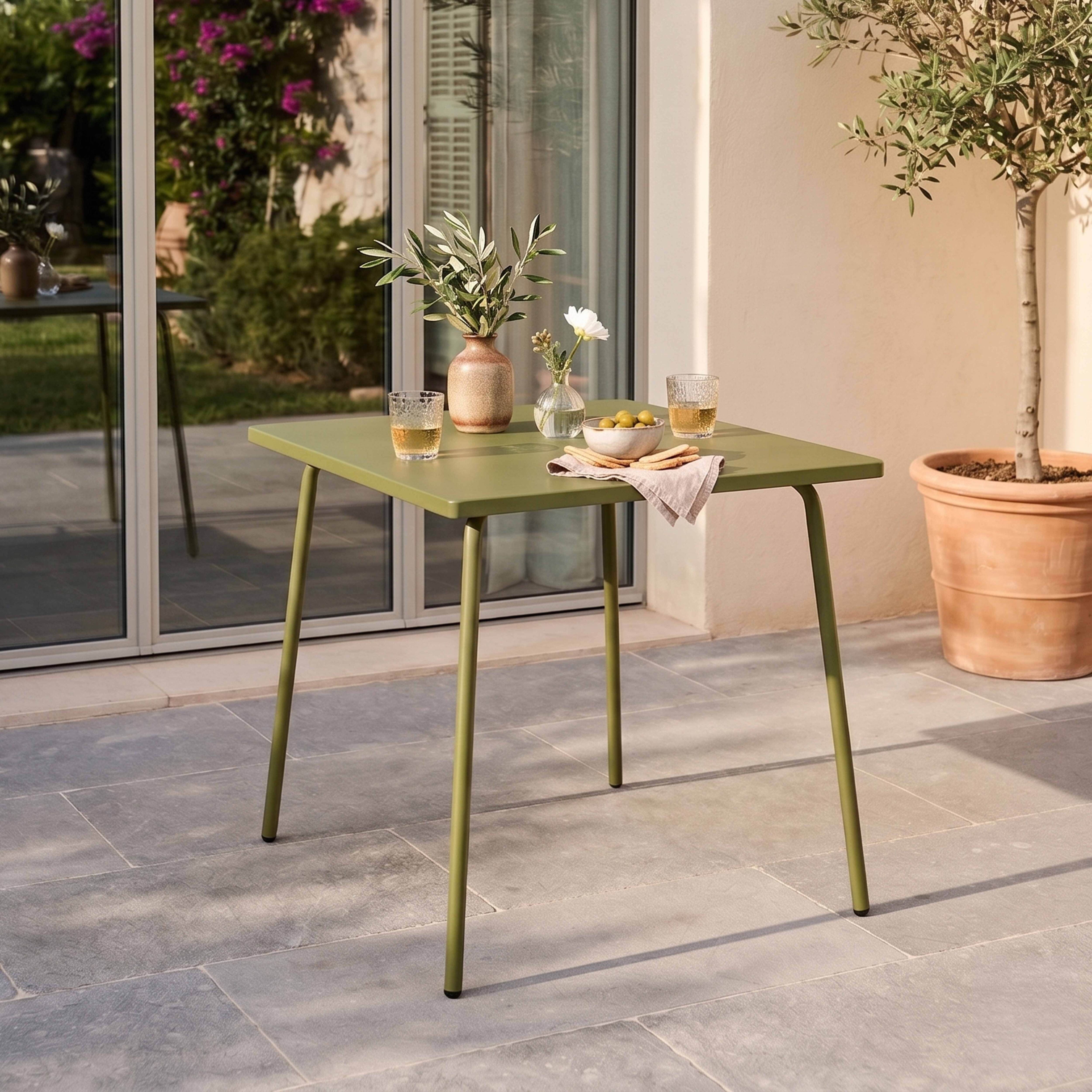 Ein grüner Gartentisch mit zwei Gläsern, einer Vase mit Olivenzweigen und einer Schale Oliven.