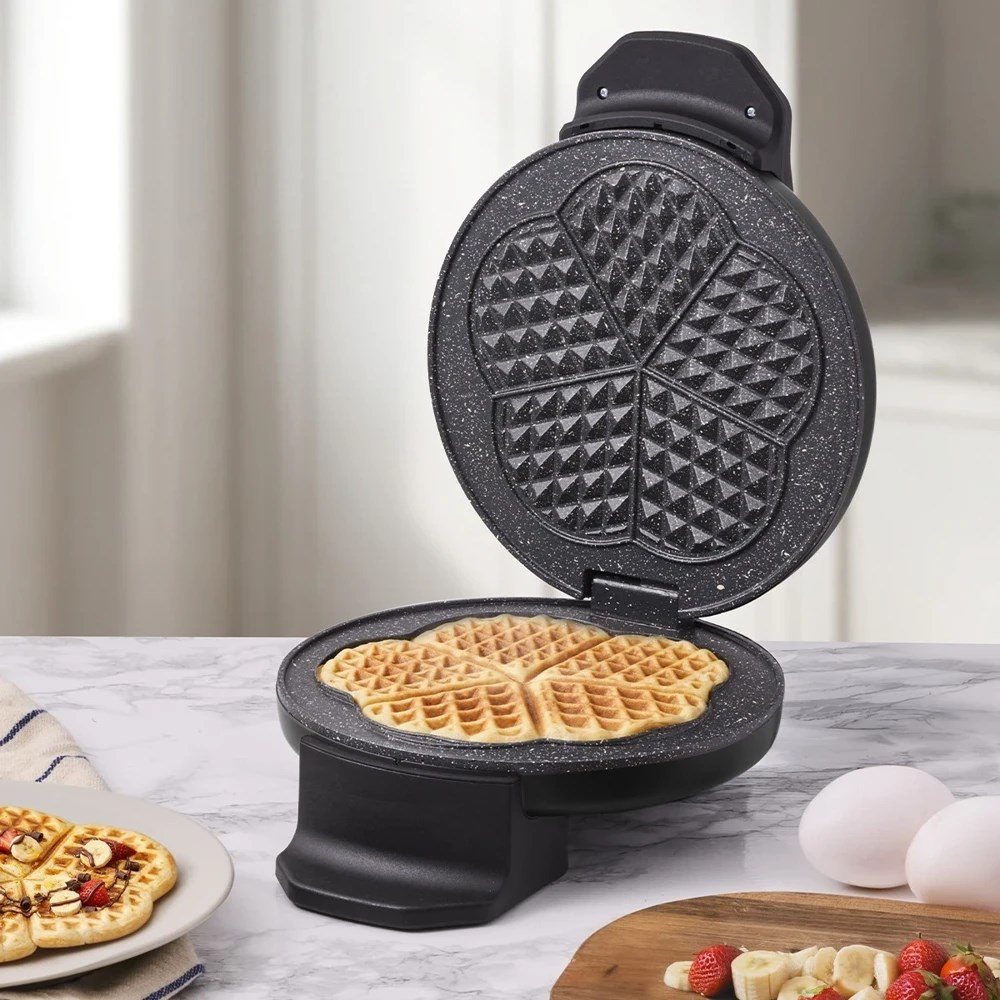 Neredeyse pişmiş bir waffle, yanında yumurta ve meyveler bulunan açık bir waffle makinesi.
