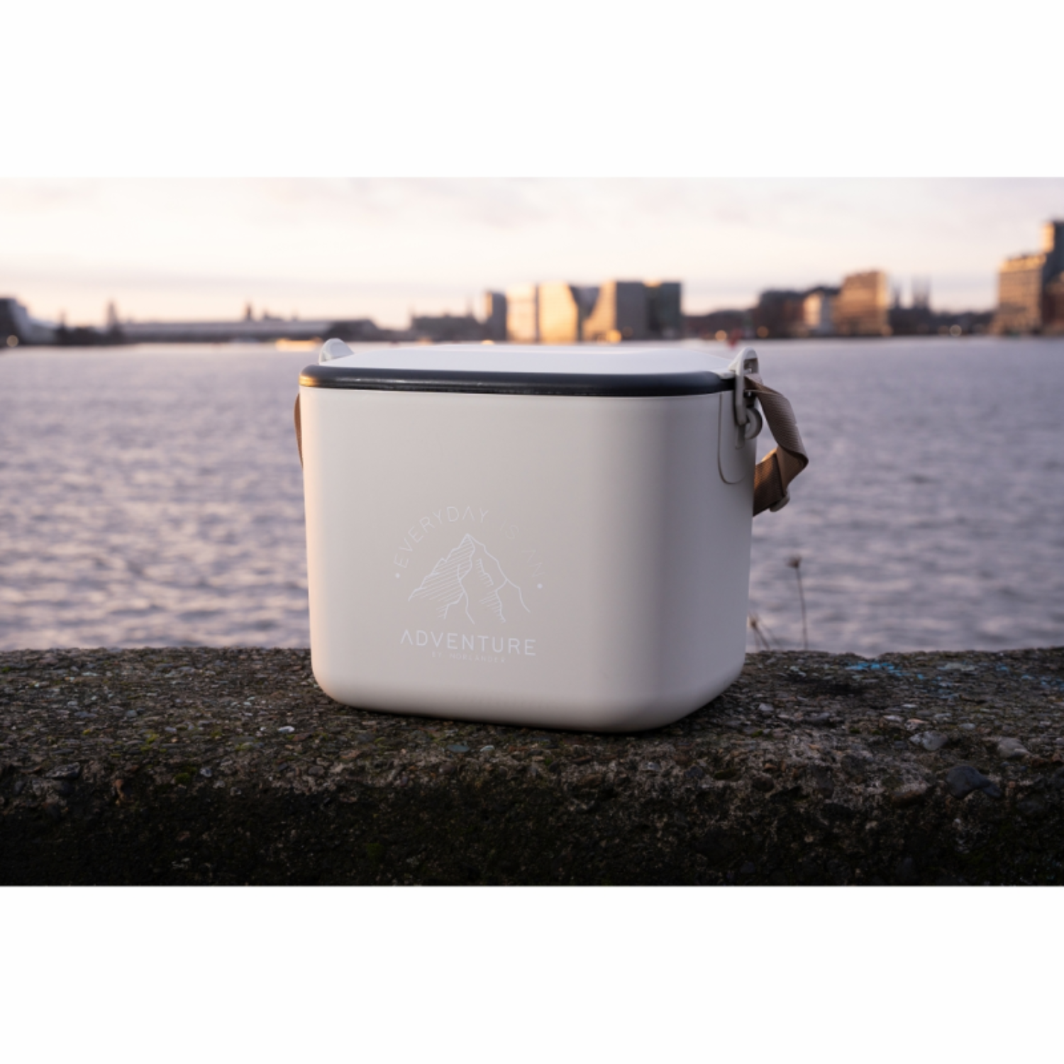 Een lichtgekleurde lunchbox staat op een betonnen richel met uitzicht op een wateroppervlak en skyline van de stad.