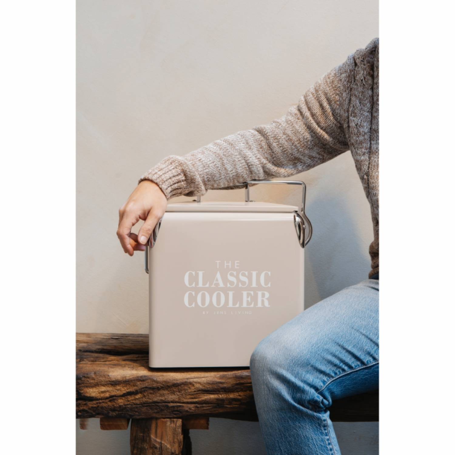 Een persoon in spijkerbroek en trui zit op een houten bankje, naast een beige koelbox.