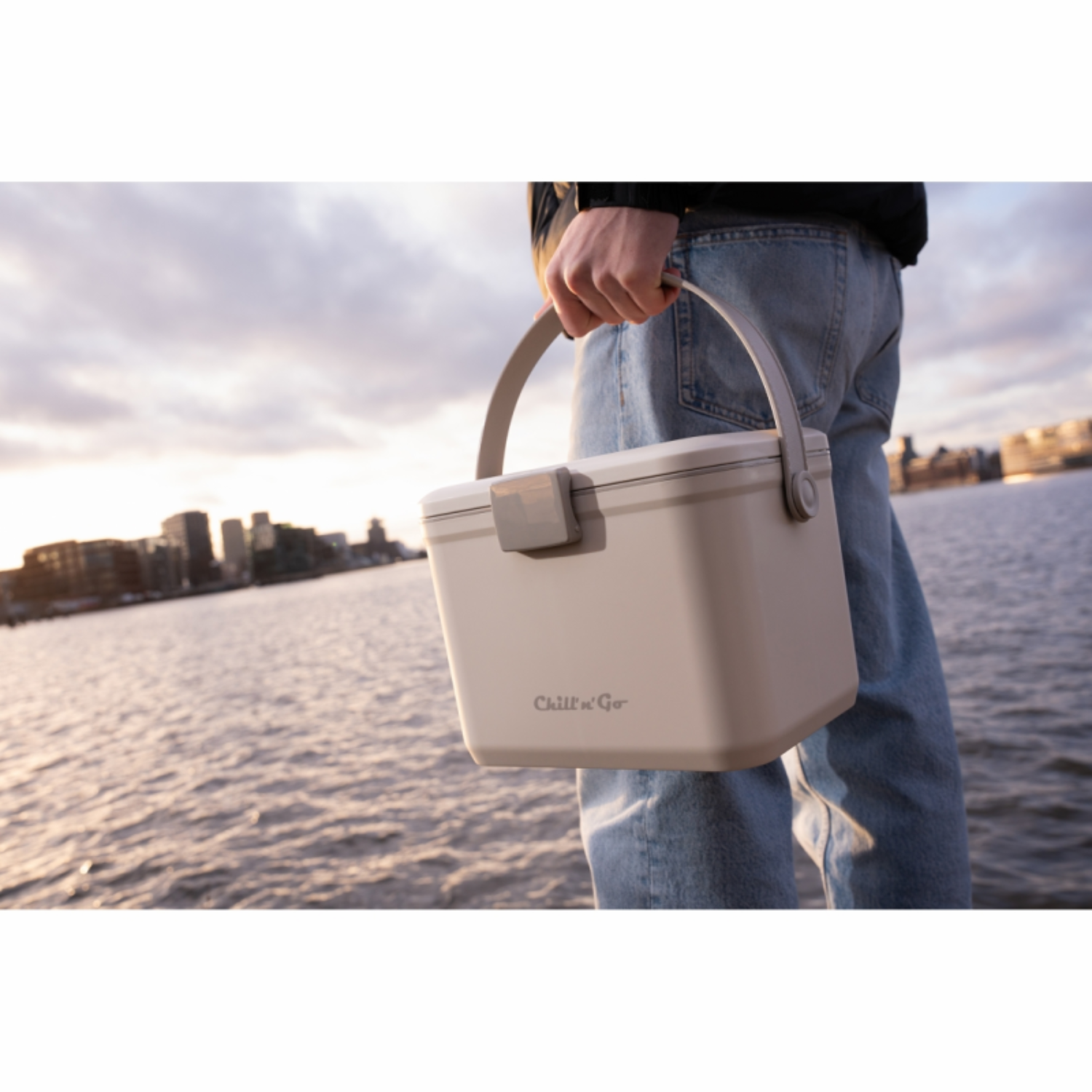 Een persoon draagt een beige koeltas met handvat aan het water.