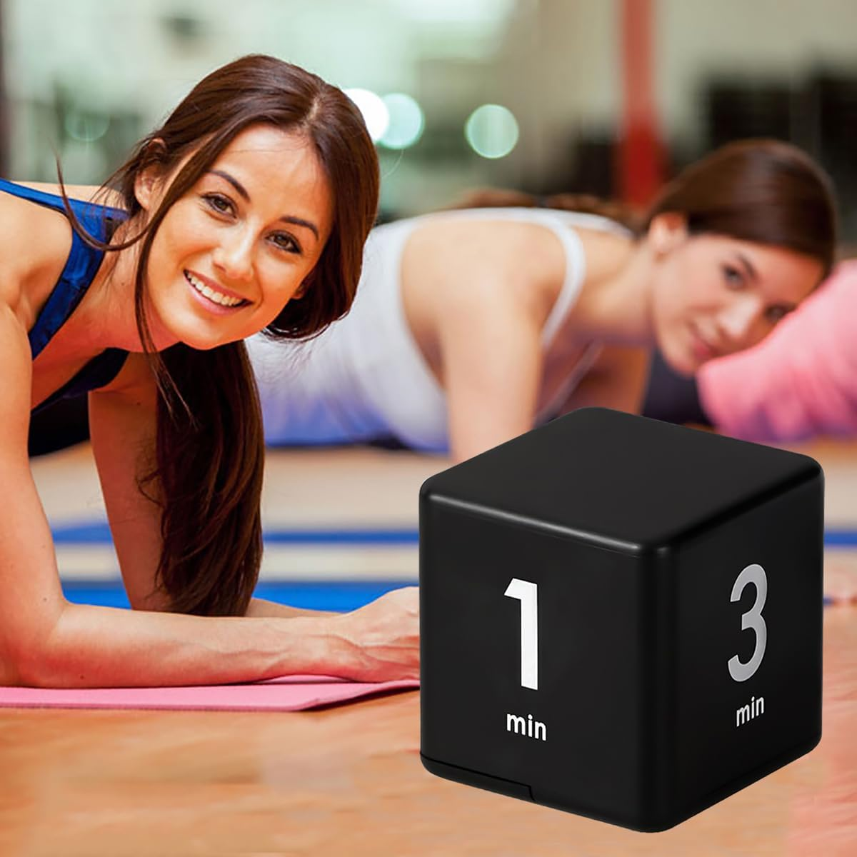 Zwei Frauen machen Plank-Übungen in einem Fitnessstudio, ein schwarzer Timer liegt vorne.