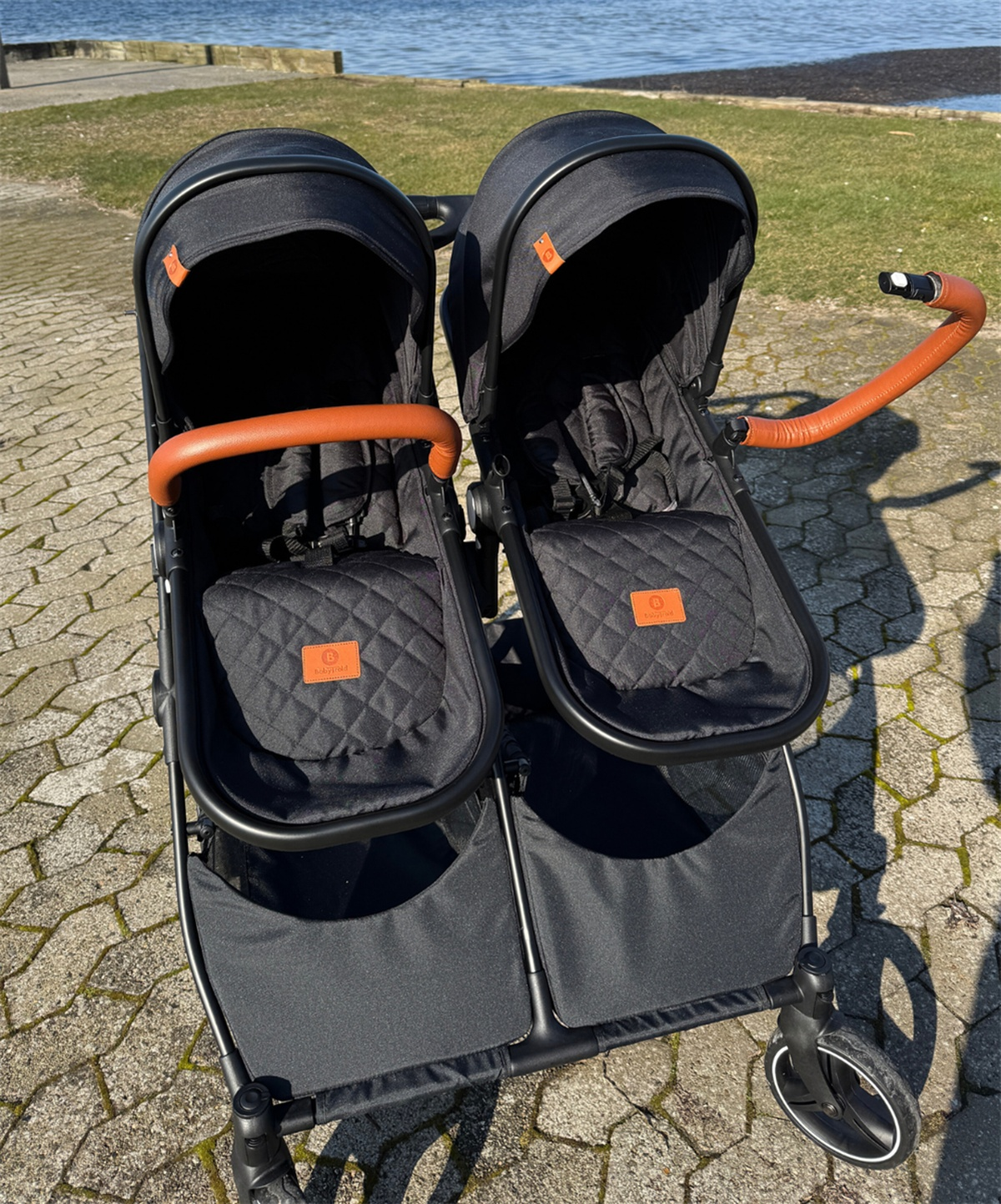 Zwei schwarze Kinderwagen nebeneinander mit braunen Griffen vor einem See.