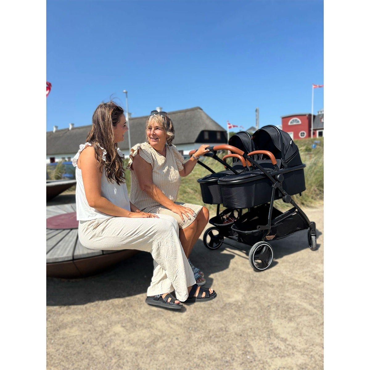 Zwei Frauen sitzen an einem sonnigen Tag auf einer Bank neben einem Doppelwagen.