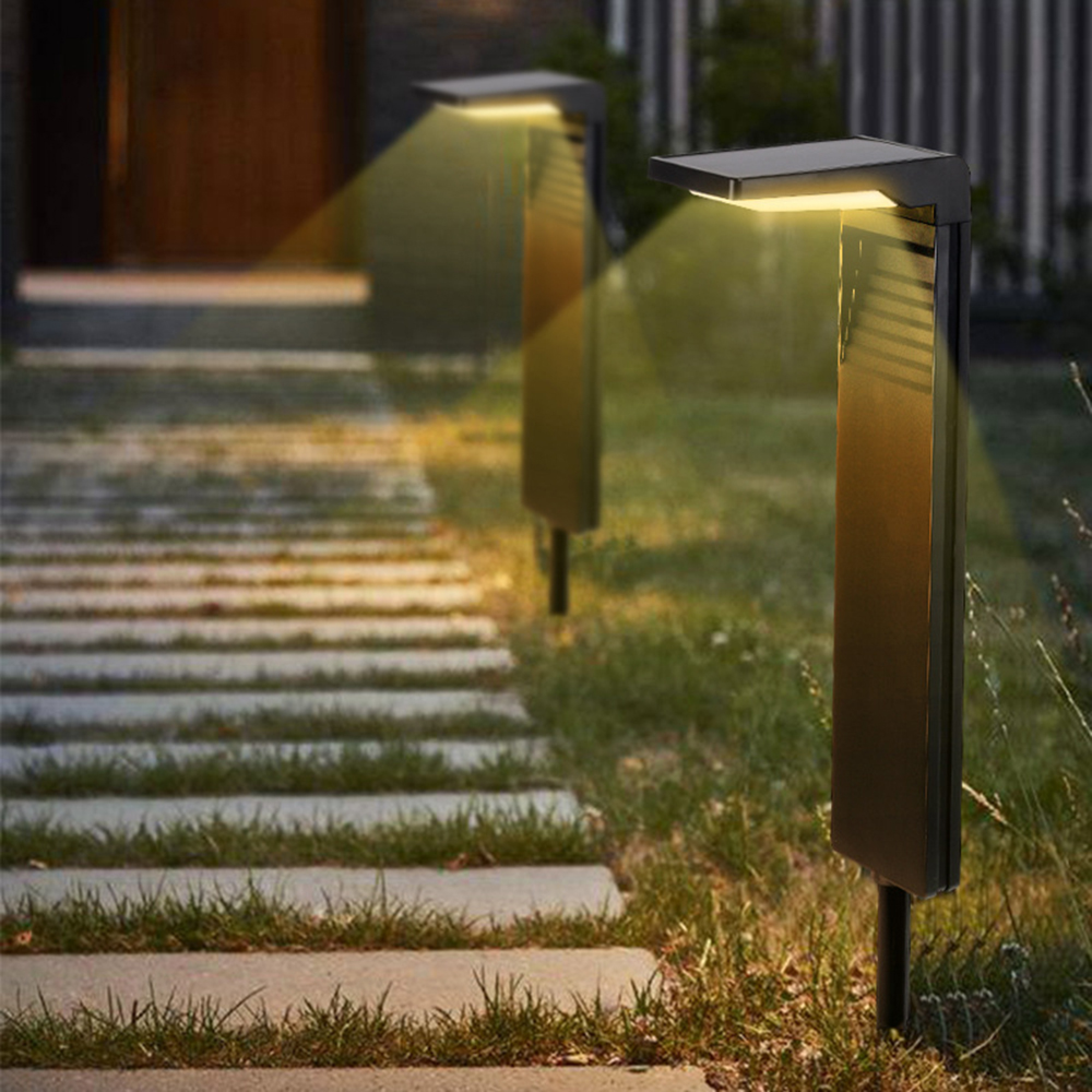 Zwei moderne Solar-Wegeleuchten beleuchten einen Steinweg durch einen grasbewachsenen Hof in der Dämmerung.