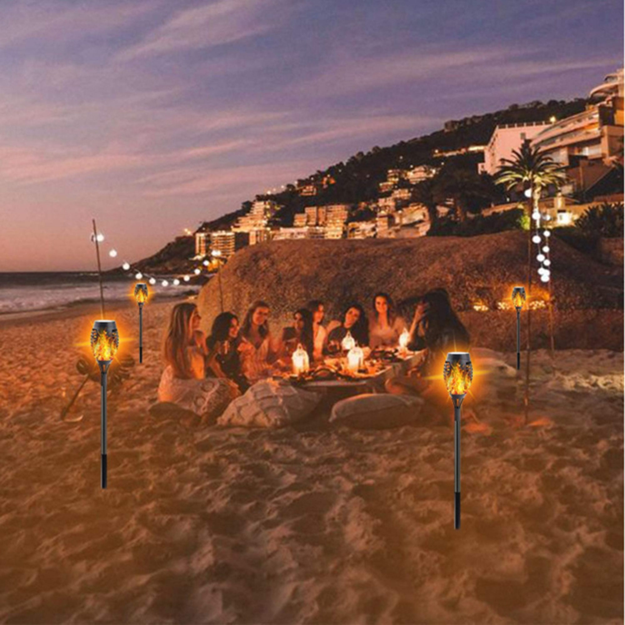 Menschen sitzen am Strand bei Fackellicht, im Hintergrund eine Stadt am Hang.