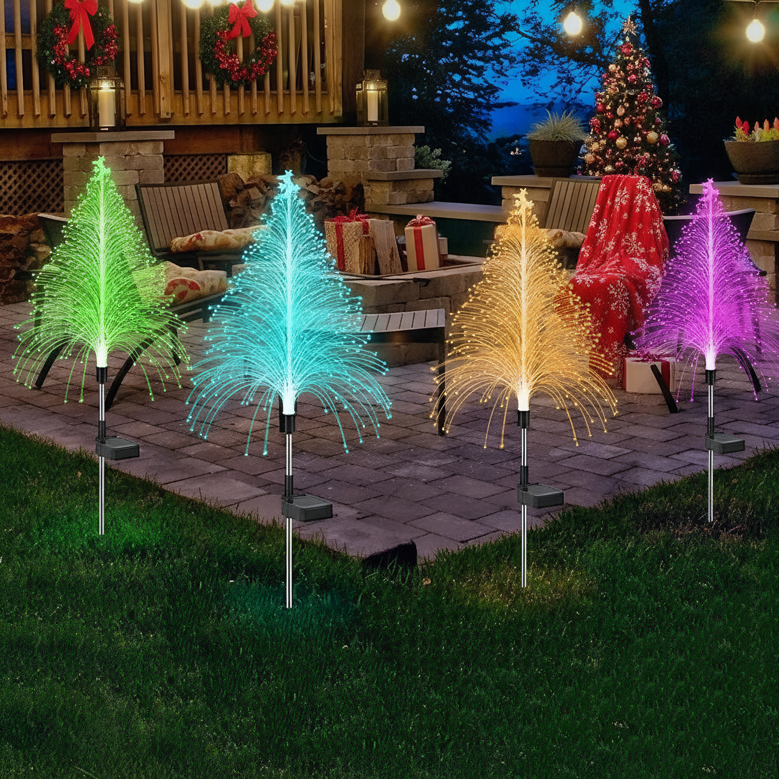 Vier leuchtende Faserbaum-Dekorationen in Grün, Blau, Orange und Lila auf einer Terrasse bei Nacht.