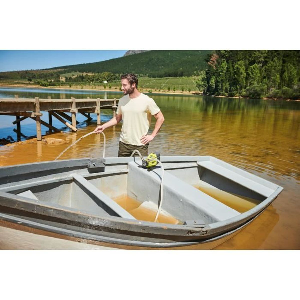 Mann pumpt Wasser aus Boot am Steg. See und Bäume im Hintergrund.