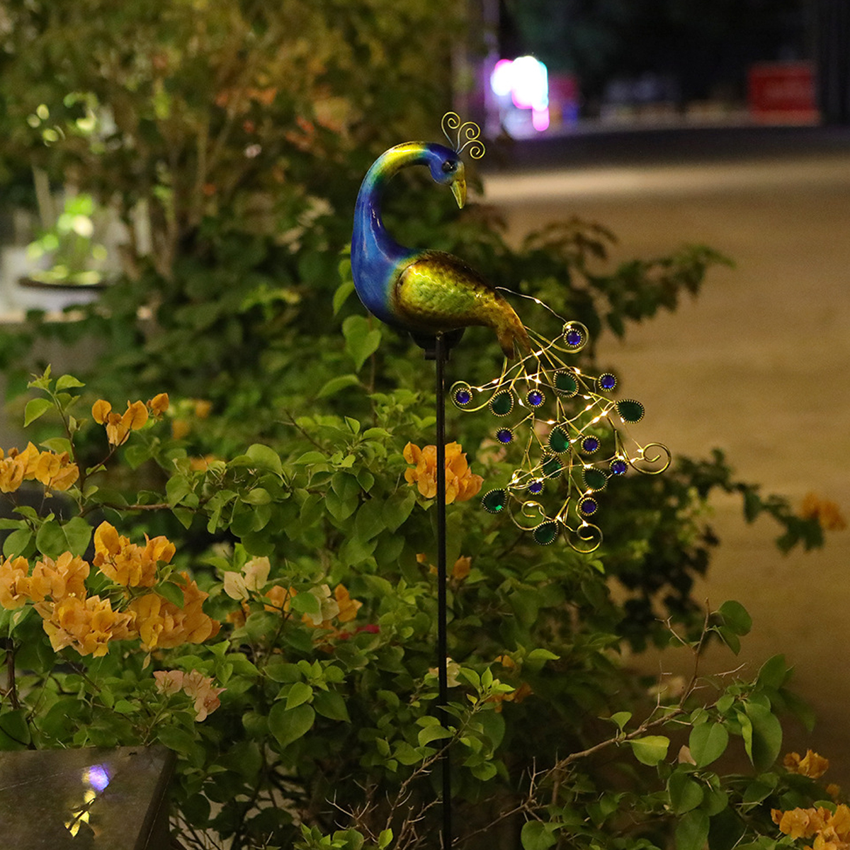 Ein metallischer Pfauengartenstecker mit Lichtern steht zwischen Blumen im Dunkeln.