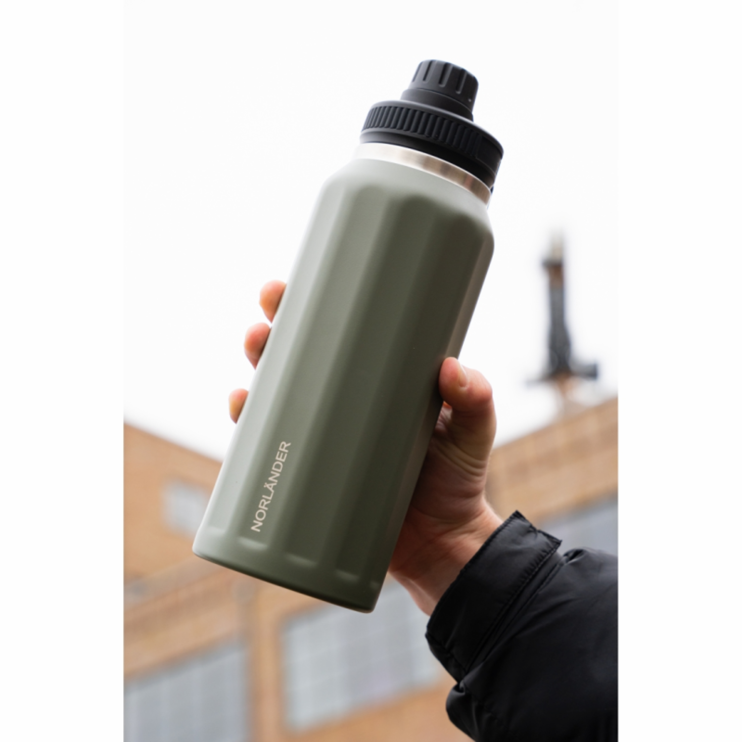 Een hand houdt een olijfgroene thermosfles met zwarte dop vast, voor wazige gebouwen.