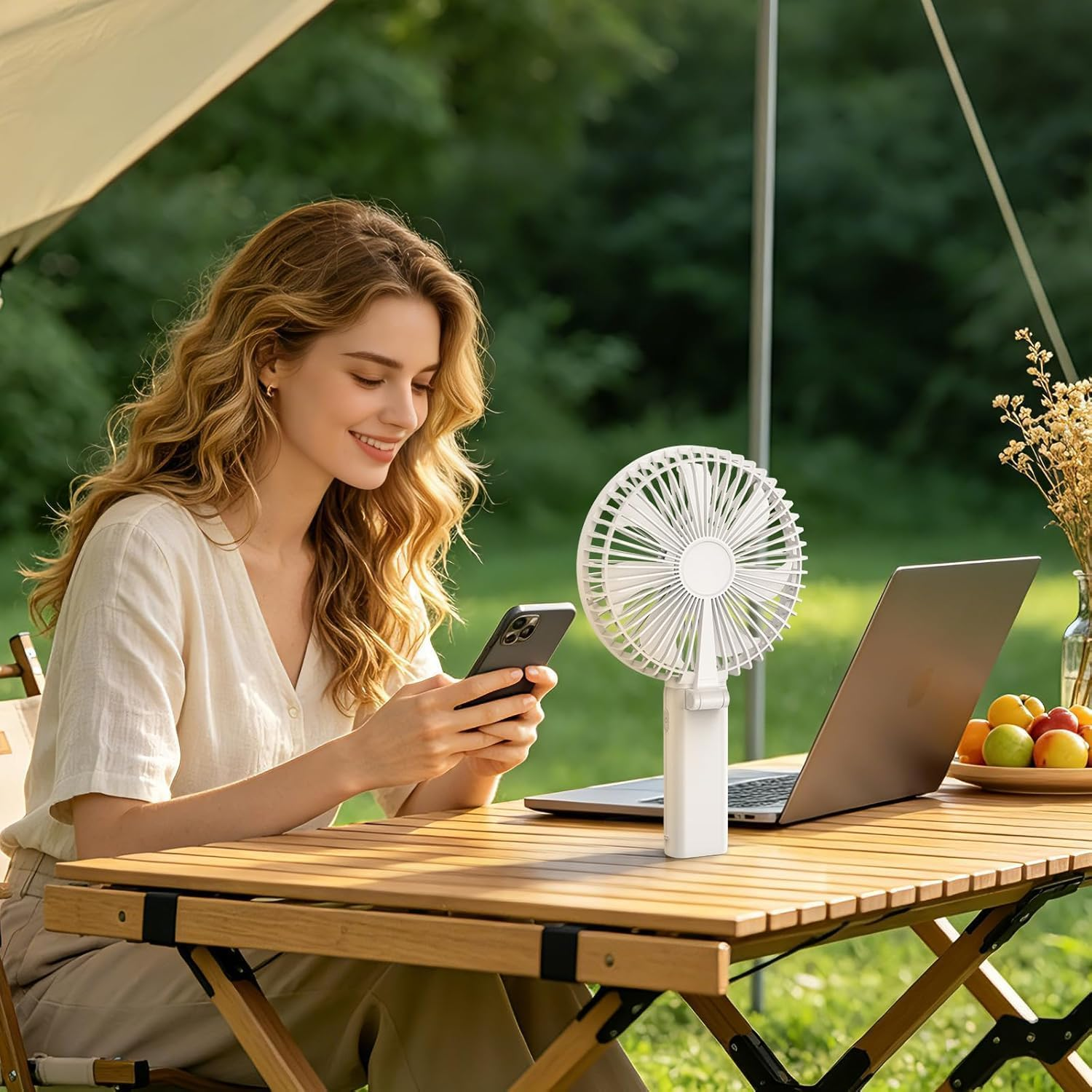 Frau am Handy am Tisch mit Laptop, Ventilator und Obst draußen.