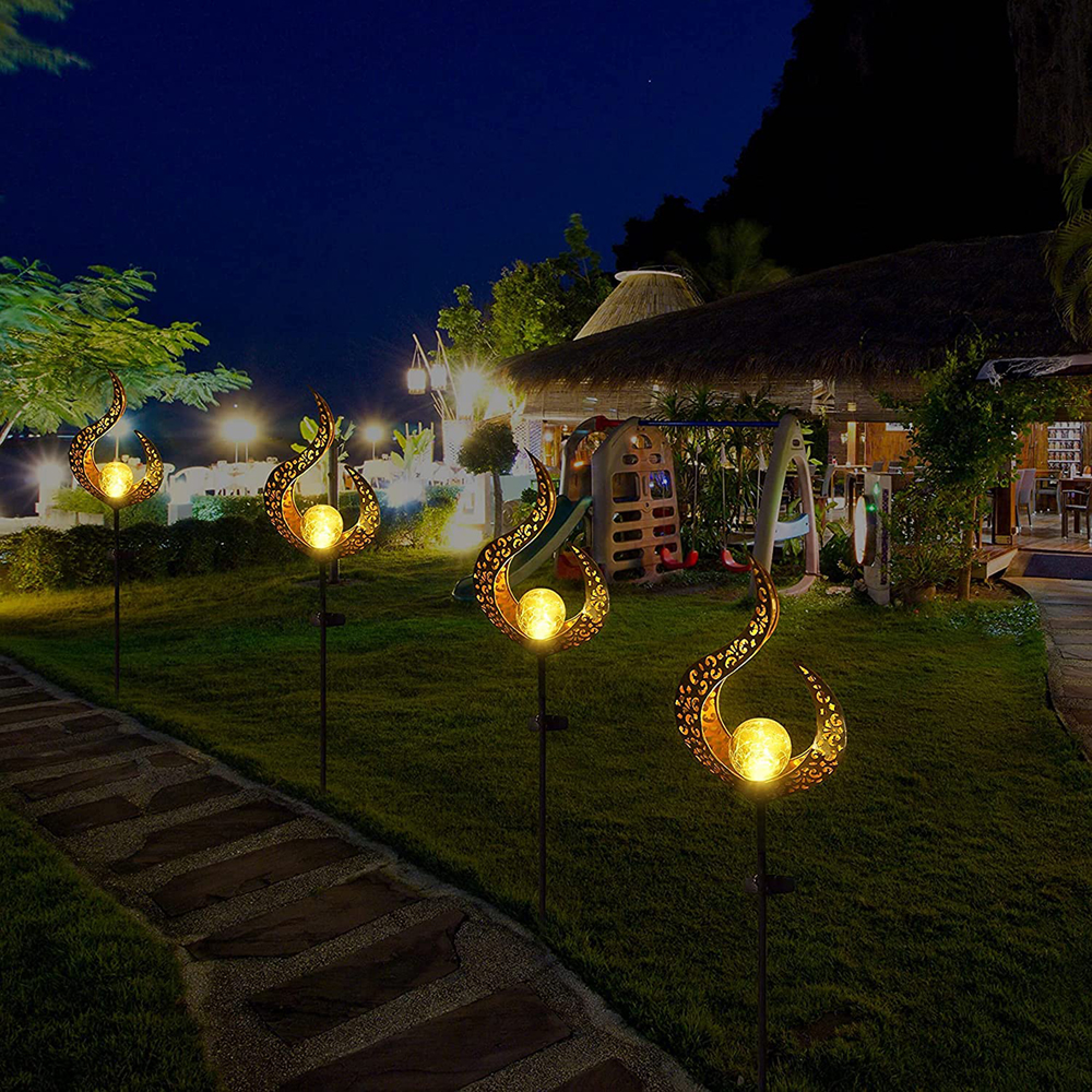 Drei leuchtende Gartenleuchten mit Flammenmuster auf einem Rasen neben einem Weg bei Nacht.