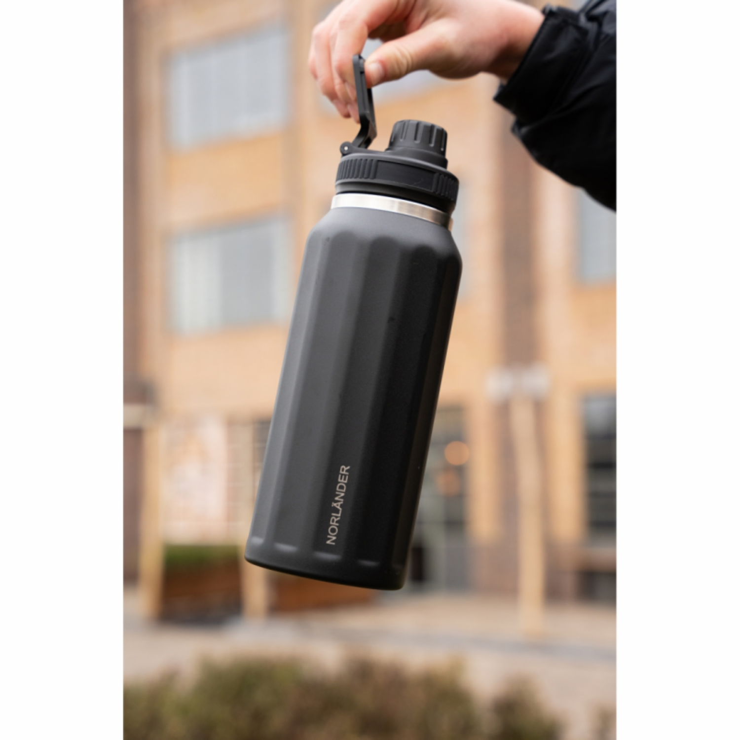 Een hand houdt een zwarte waterfles met NORLÄNDER-logo voor een wazig gebouw.