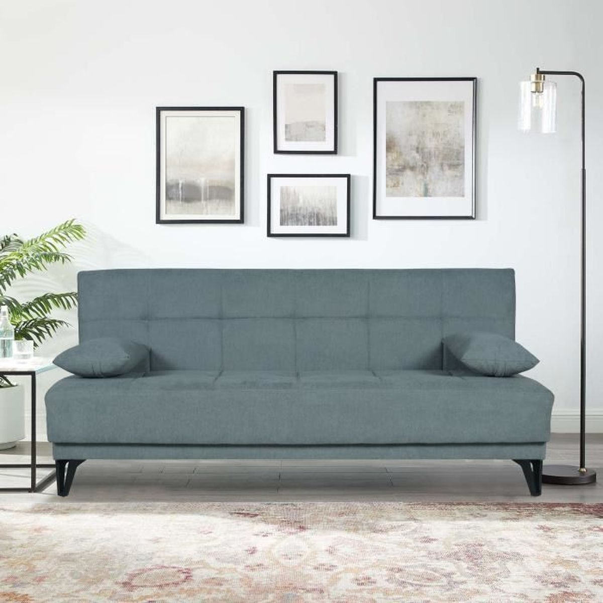 Ein graublaues Schlafsofa mit zwei Kissen steht auf einem Teppich. Vier Bilder hängen an der Wand.
