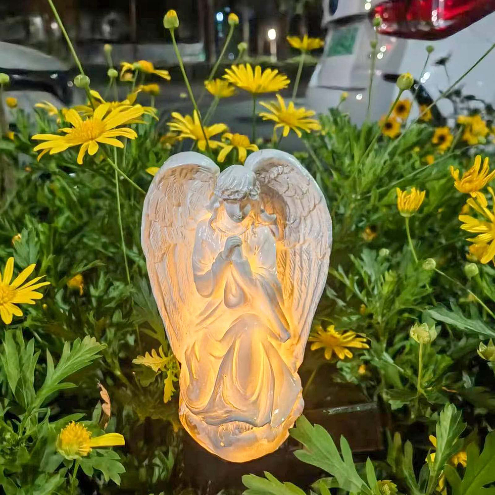 Ein beleuchteter Engelsengel steht inmitten gelber Blumen und grüner Blätter.