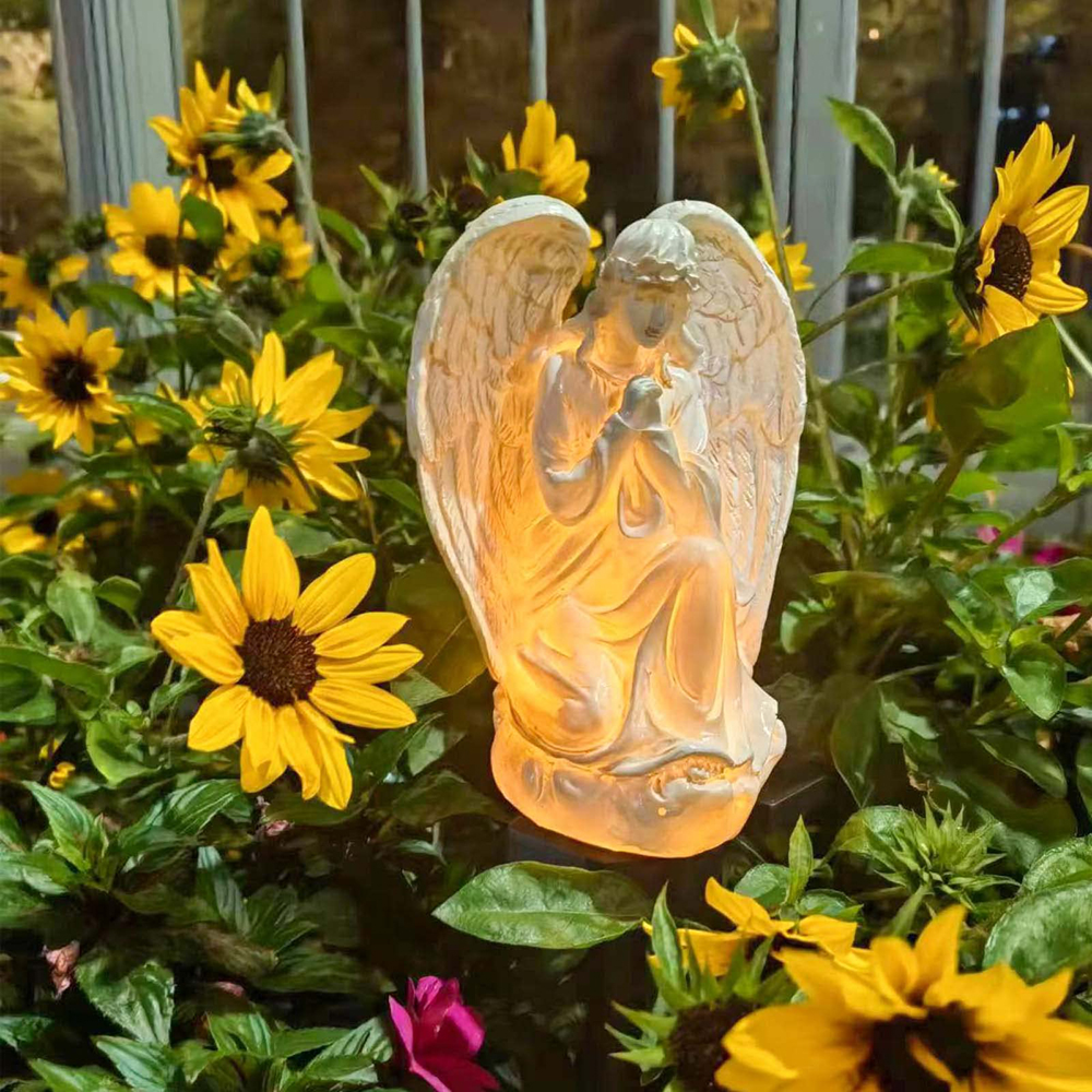 Eine leuchtende Engelsstatue kniet inmitten von Sonnenblumen und grünem Laub.