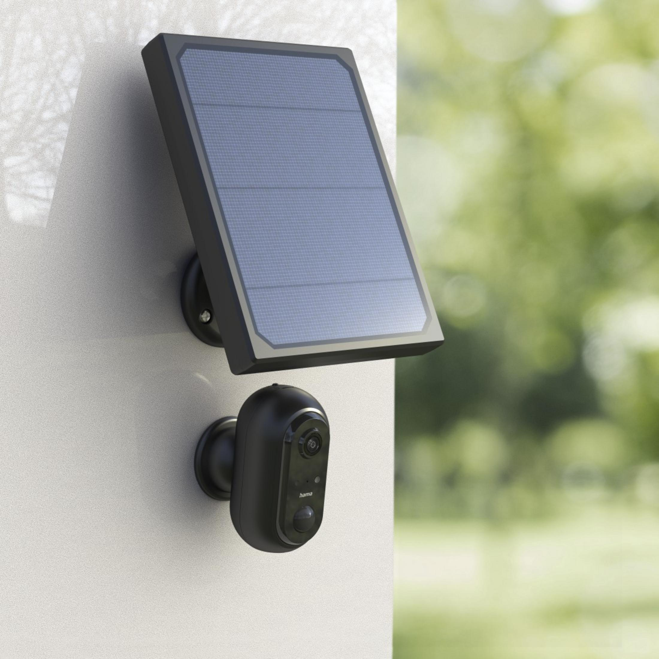 Schwarze Überwachungskamera und Solarpanel an weißer Wand, grüner Hintergrund.