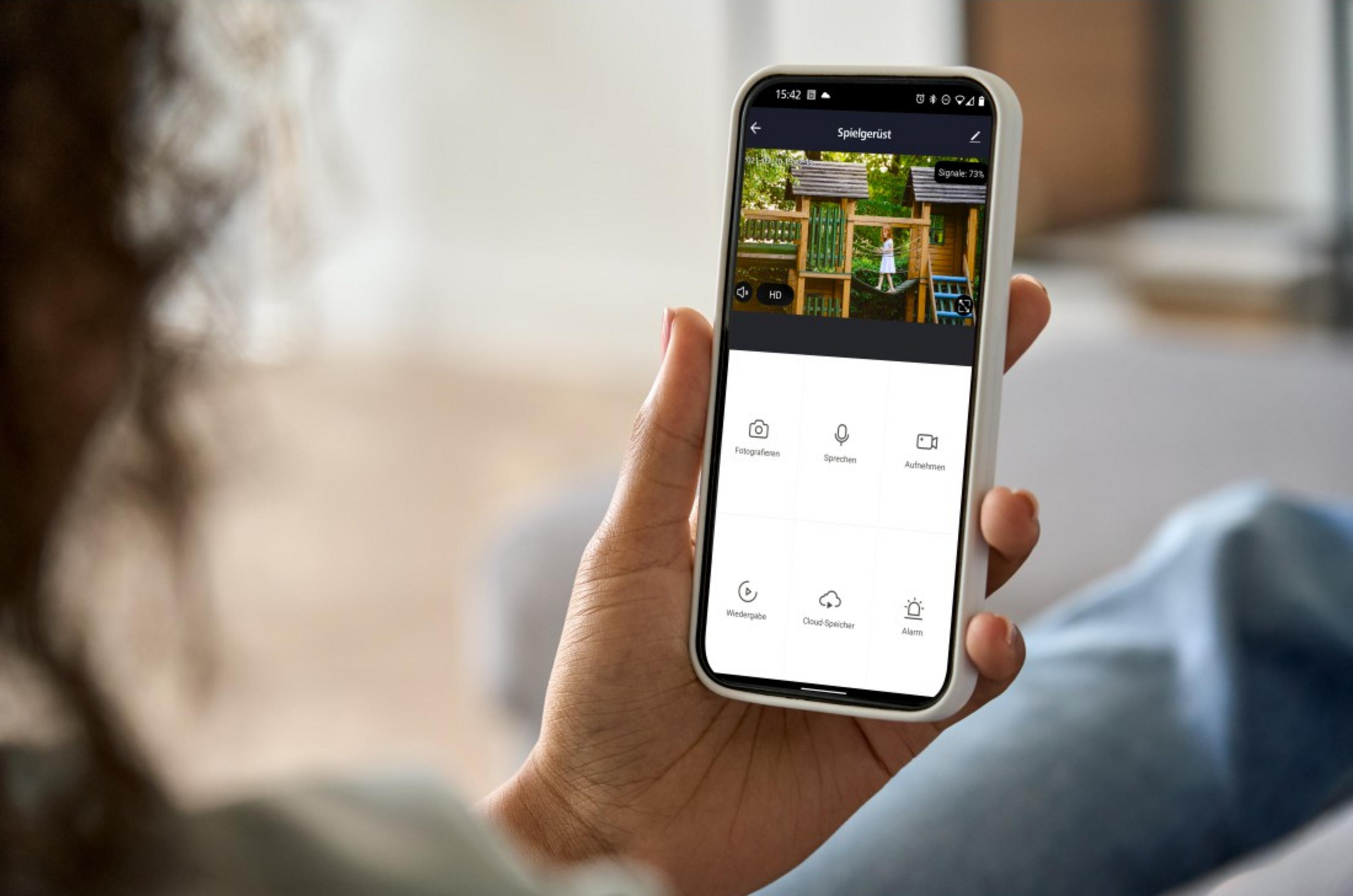 Person hält ein Smartphone, das eine Überwachungskamera-Ansicht eines Spielplatzes zeigt.