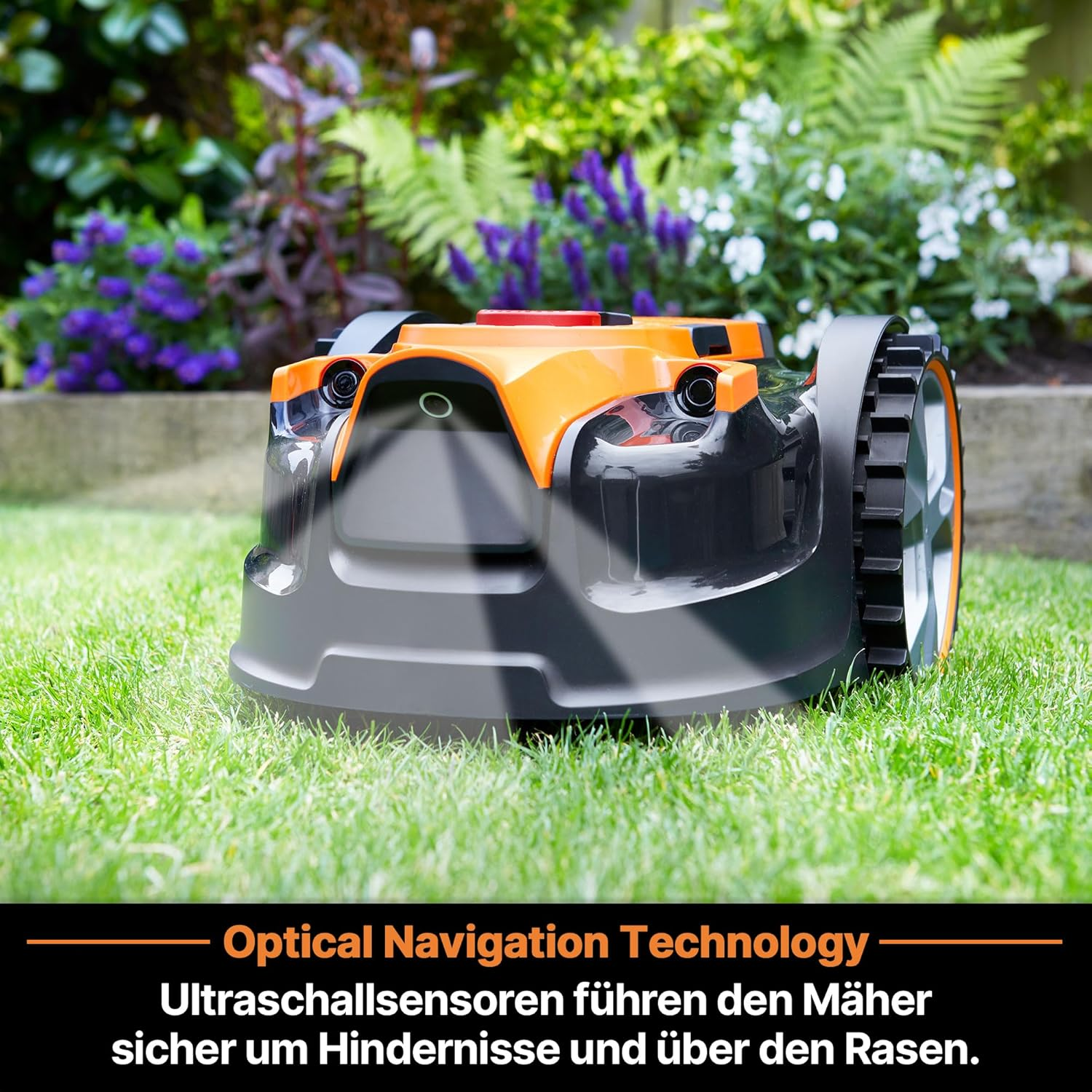 Ein orange-schwarzer Mähroboter steht auf einem grünen Rasen vor blühenden Pflanzen.