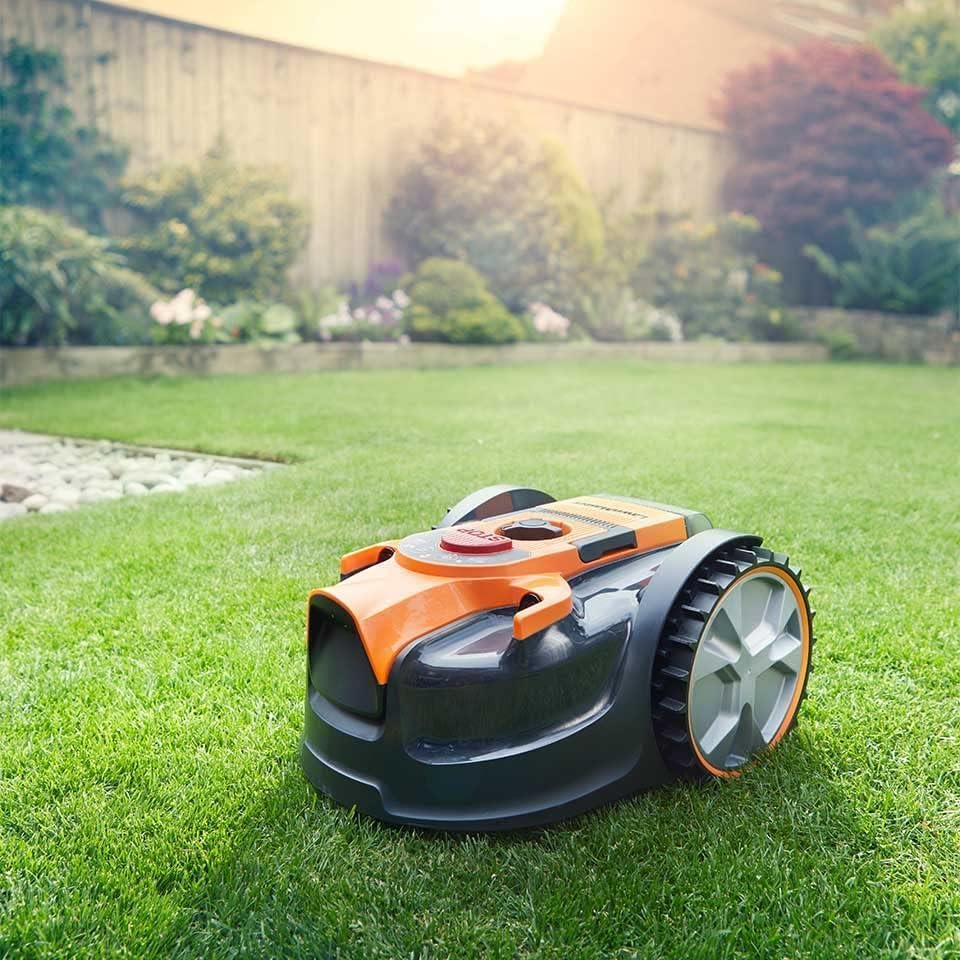 Ein Mähroboter steht auf einem grünen Rasen mit einem Zaun und Bäumen im Hintergrund.