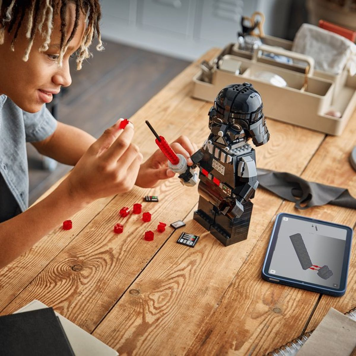 Bir kişi ahşap bir masada kırmızı parçalarla siyah bir Lego Darth Vader inşa ediyor.