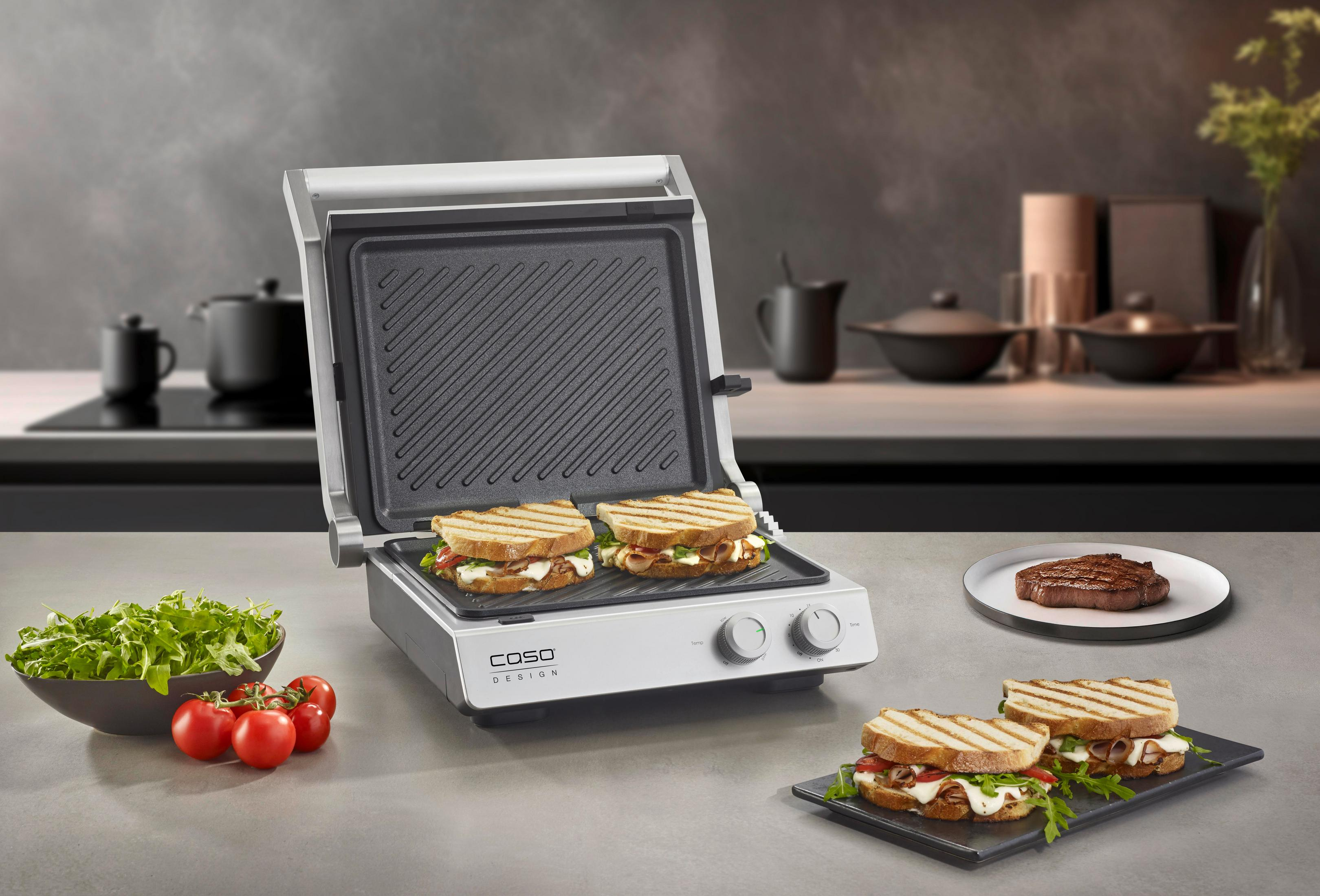 Una griglia a contatto aperta con due panini, contorni e una bistecca su un piatto.