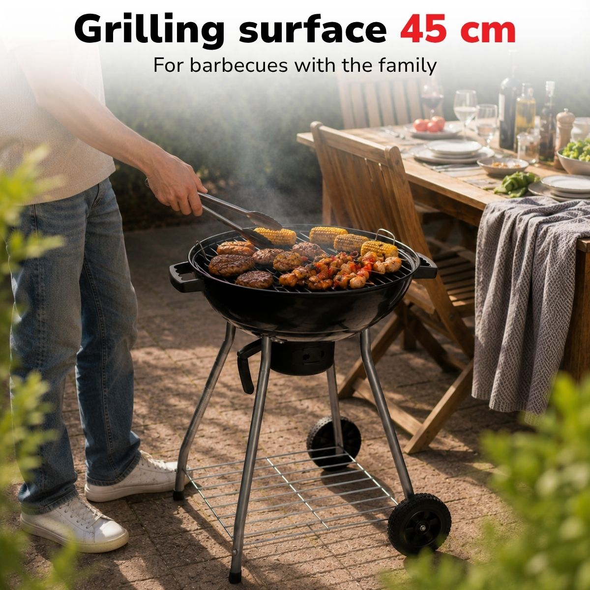 A person grills burgers and corn on a round barbecue with a table in the background.