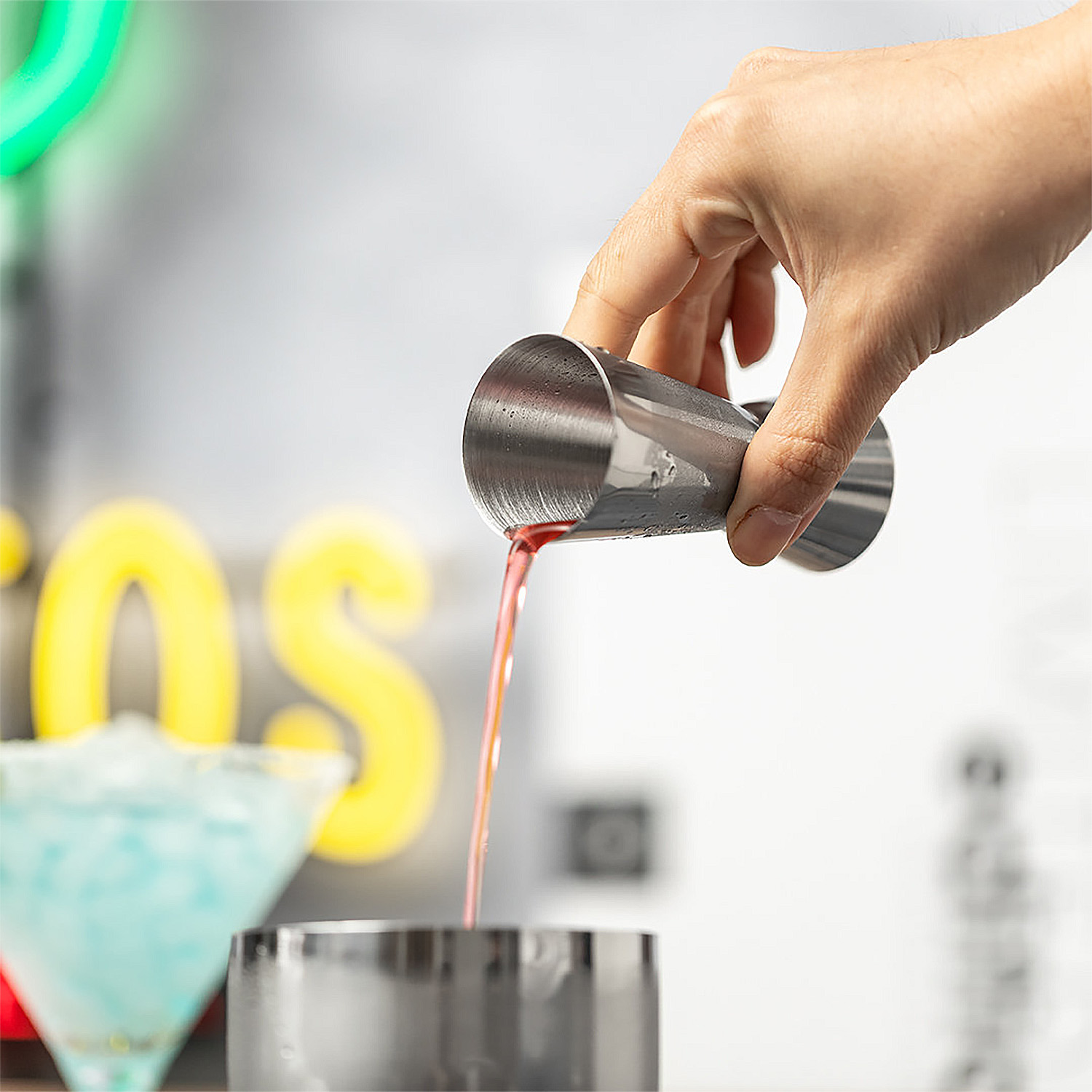 Una mano versa liquido rosso da un jigger in uno shaker per cocktail. Un margarita blu sfocato a sinistra.