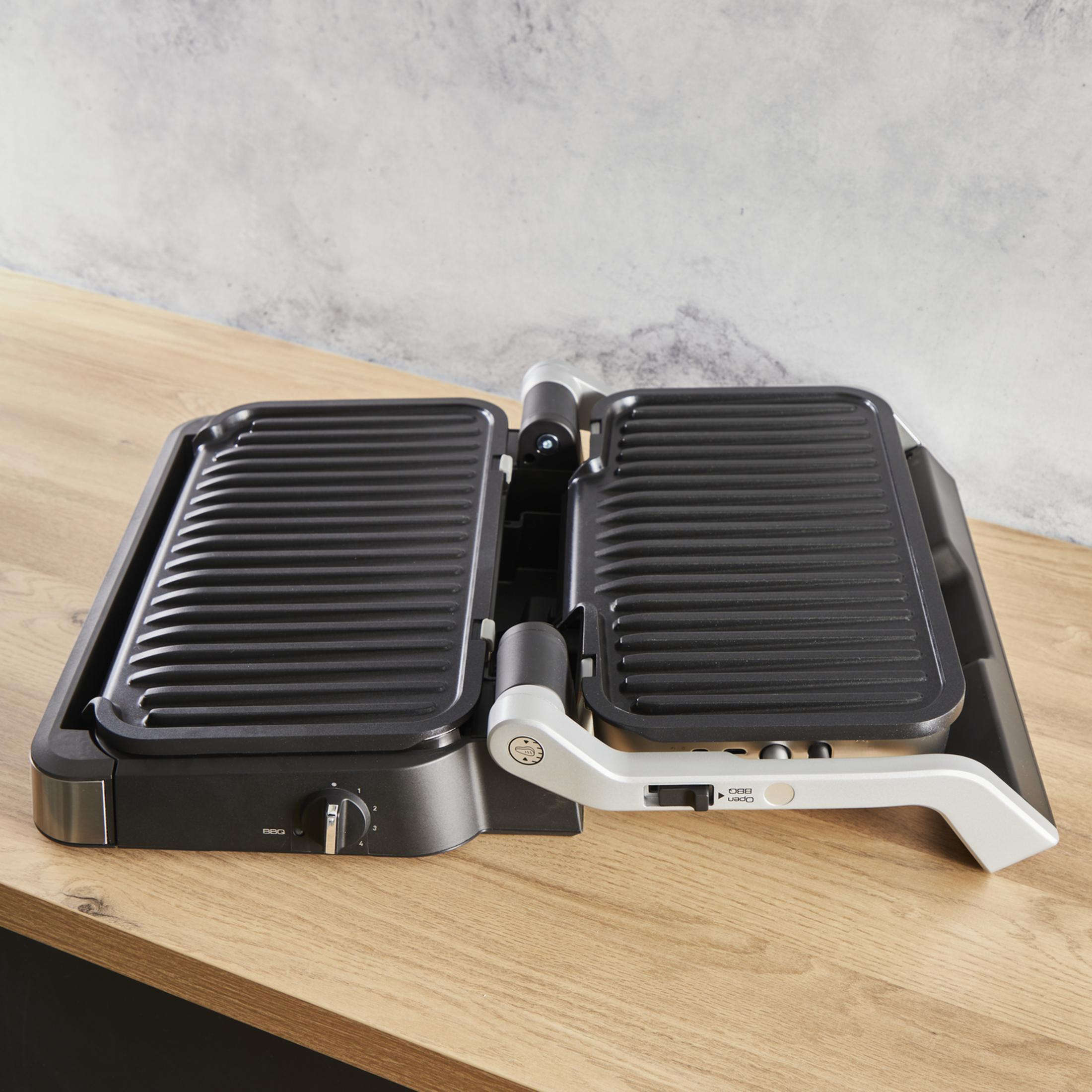 Ein offener, schwarzer Kontaktgrill mit gerippten Platten auf einem Holztisch.
