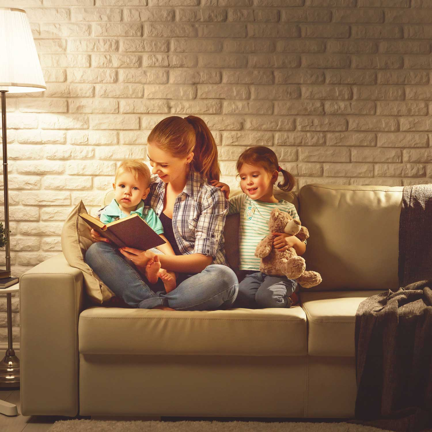 Una madre legge un libro ai suoi figli sul divano, illuminata da una lampada da terra.