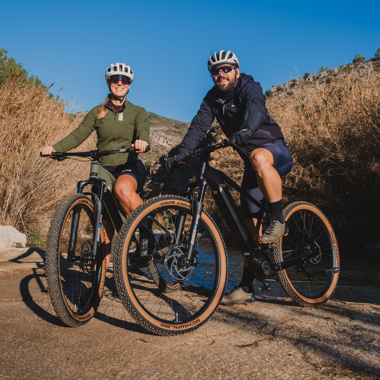 Zwei Radfahrer mit Helmen und Brillen auf E-Bikes, im Freien, mit Bäumen und Bergen im Hintergrund.