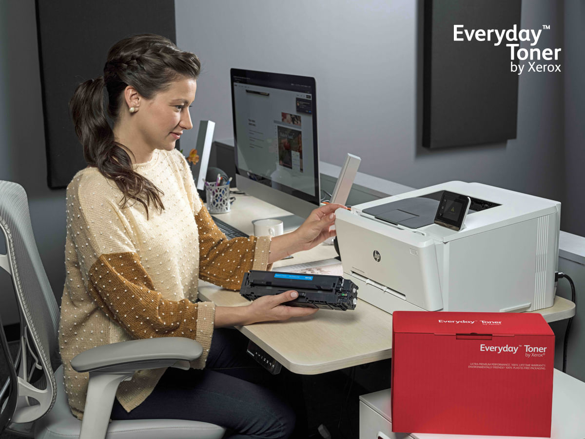 Een vrouw zit aan een bureau en houdt een tonercartridge vast naast een printer.