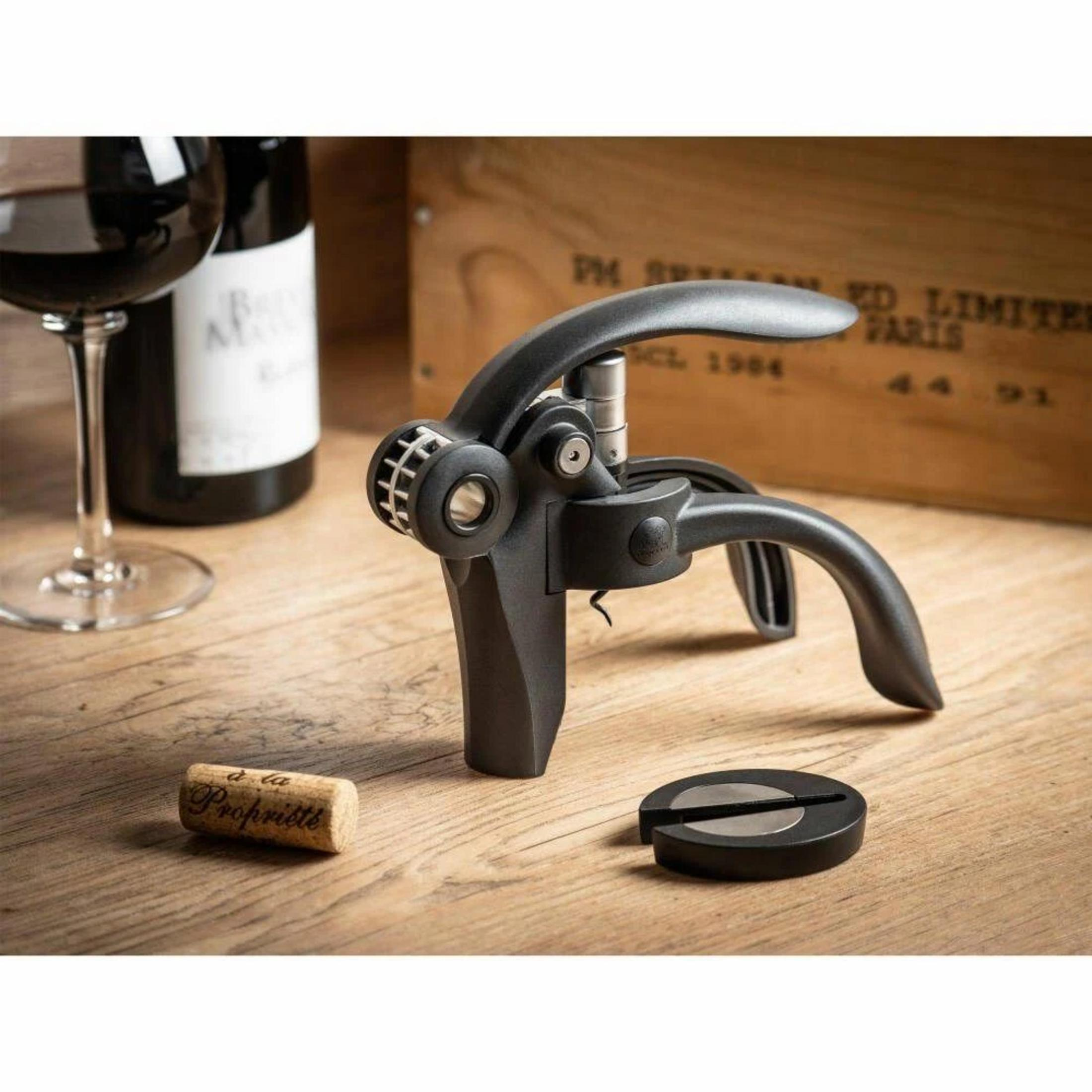 Un cavatappi nero con manici, accanto a un bicchiere di vino rosso e una bottiglia.