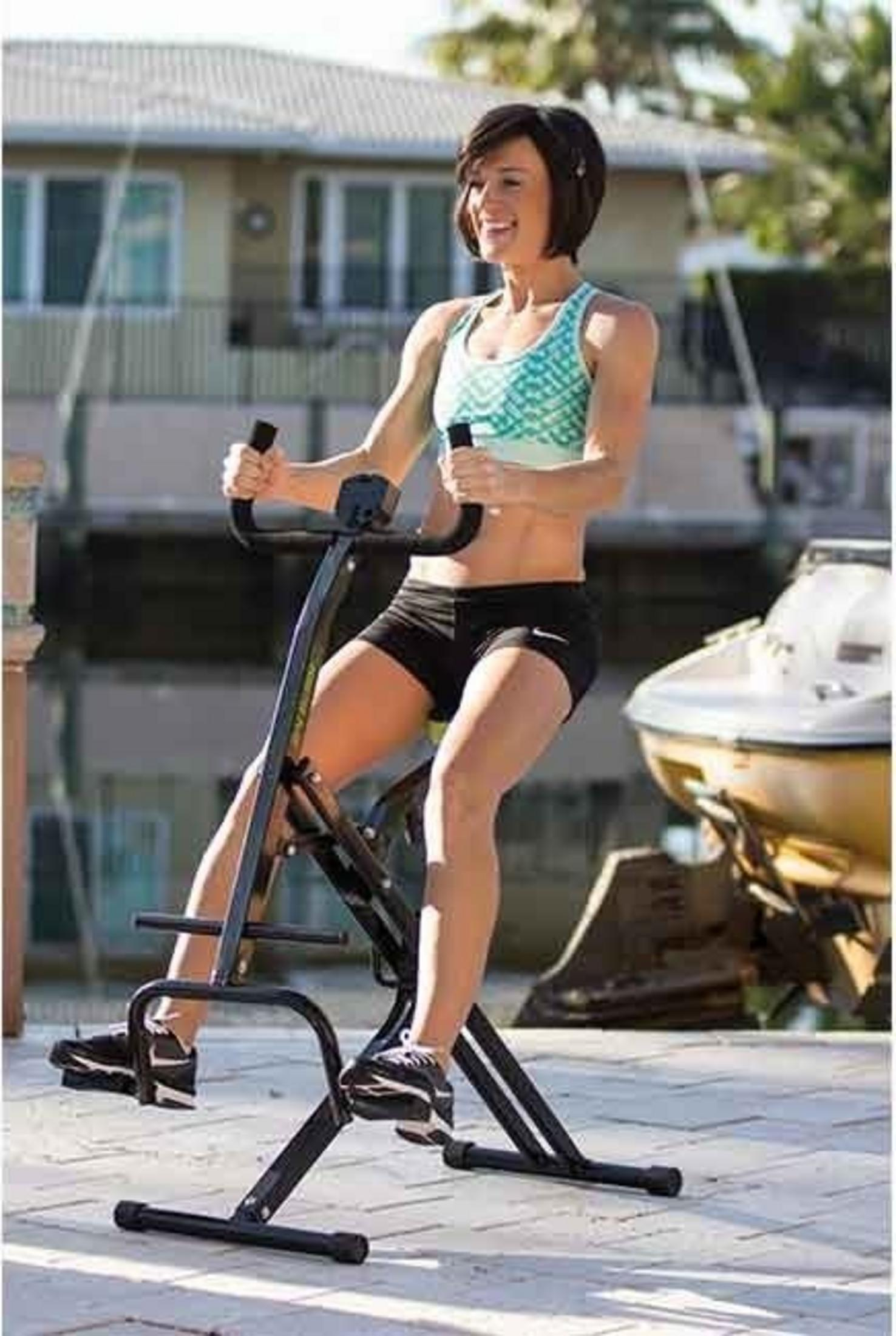 Eine Frau trainiert auf einem Heimtrainer im Freien, mit einem Boot und einem Haus im Hintergrund.