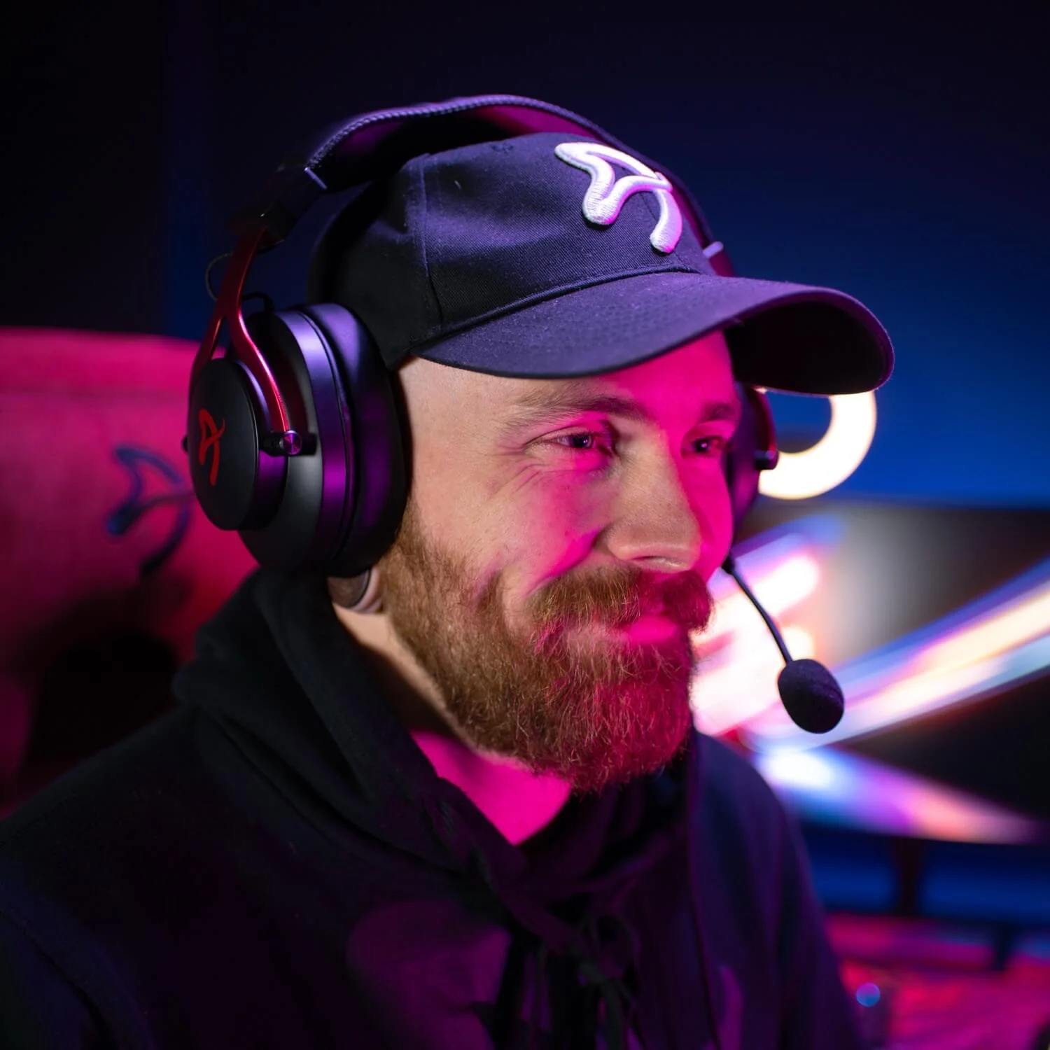 Un uomo con cappellino nero e cuffie con microfono, illuminato da luci rosa e blu.