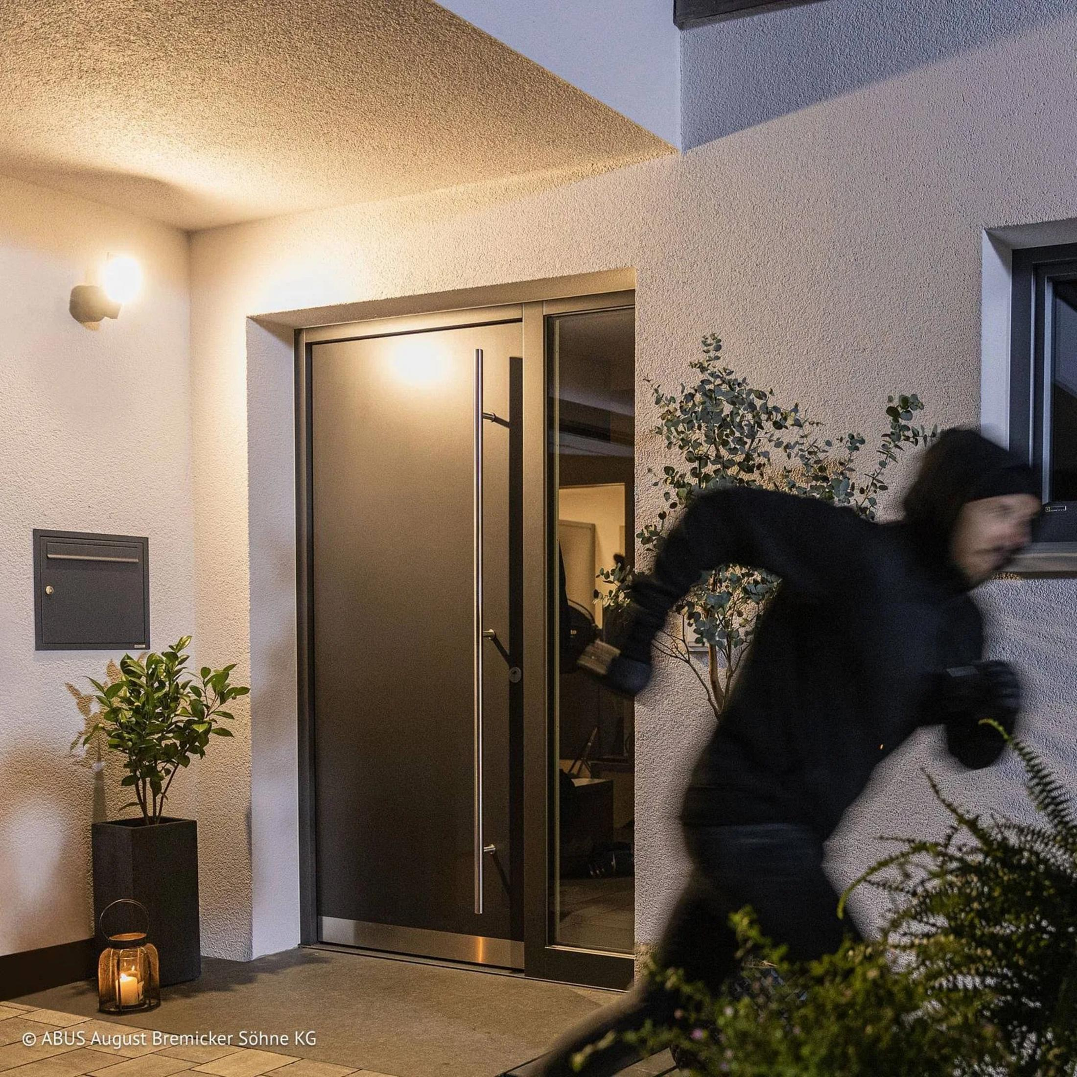 Ein Mann in dunkler Kleidung rennt über eine beleuchtete Veranda vor einer modernen Tür.