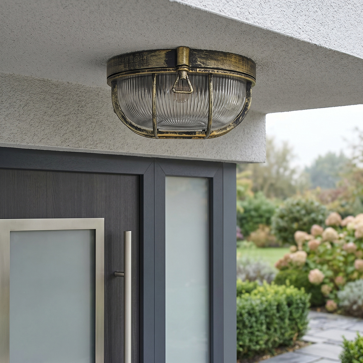Außenleuchte mit Glas und Metallkäfig über einer dunklen Tür, Garten im Hintergrund.